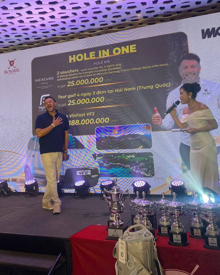 On stage at the prize dinner, Mario stands with his eyes closed and his hand on his heart while the host speaks into a microphone.
