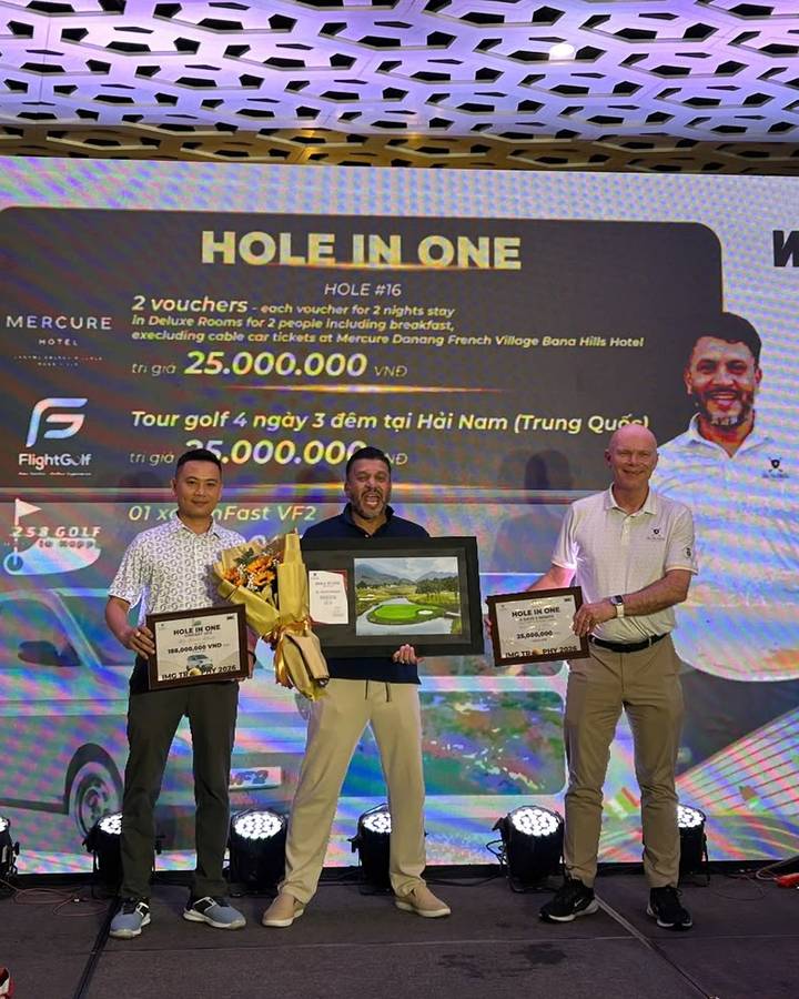 Mario on stage, shouting with joy, holding a bouquet and his framed certificate, between two presenters holding prize boards.