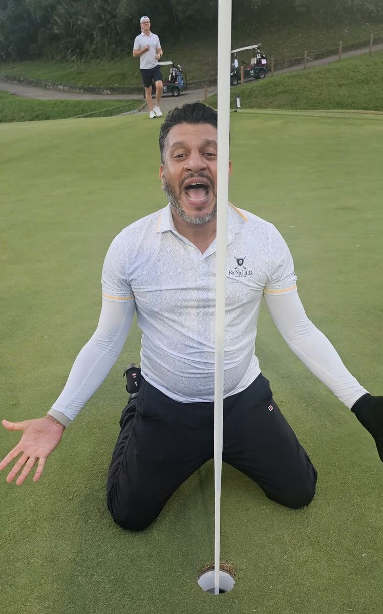 Mario on his knees on the green beside the hole, arms spread and mouth wide open, the flagstick running straight down the middle of his face. Behind him a friend is jogging over.