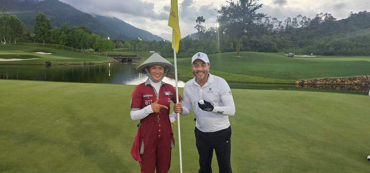 Mario beside the flag on the 16th green, holding the ball in his gloved hand. Next to him his caddie, in a conical hat and bib number 072, points at him and smiles. Mountains and a lake behind.